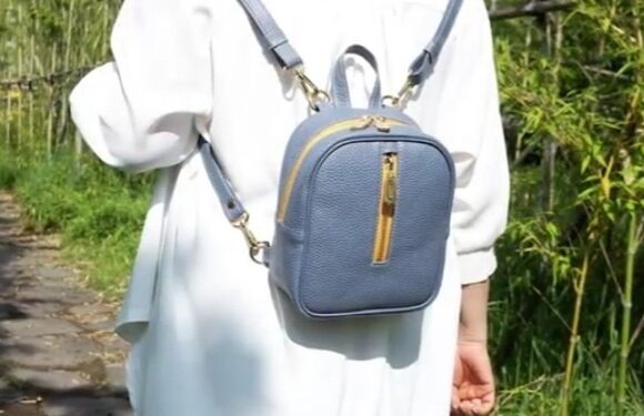 Человек в маленьком синем кожаном рюкзачке с золотыми застежками-молниями и фурнитурой идет по каменной дорожке, окруженной зеленью.